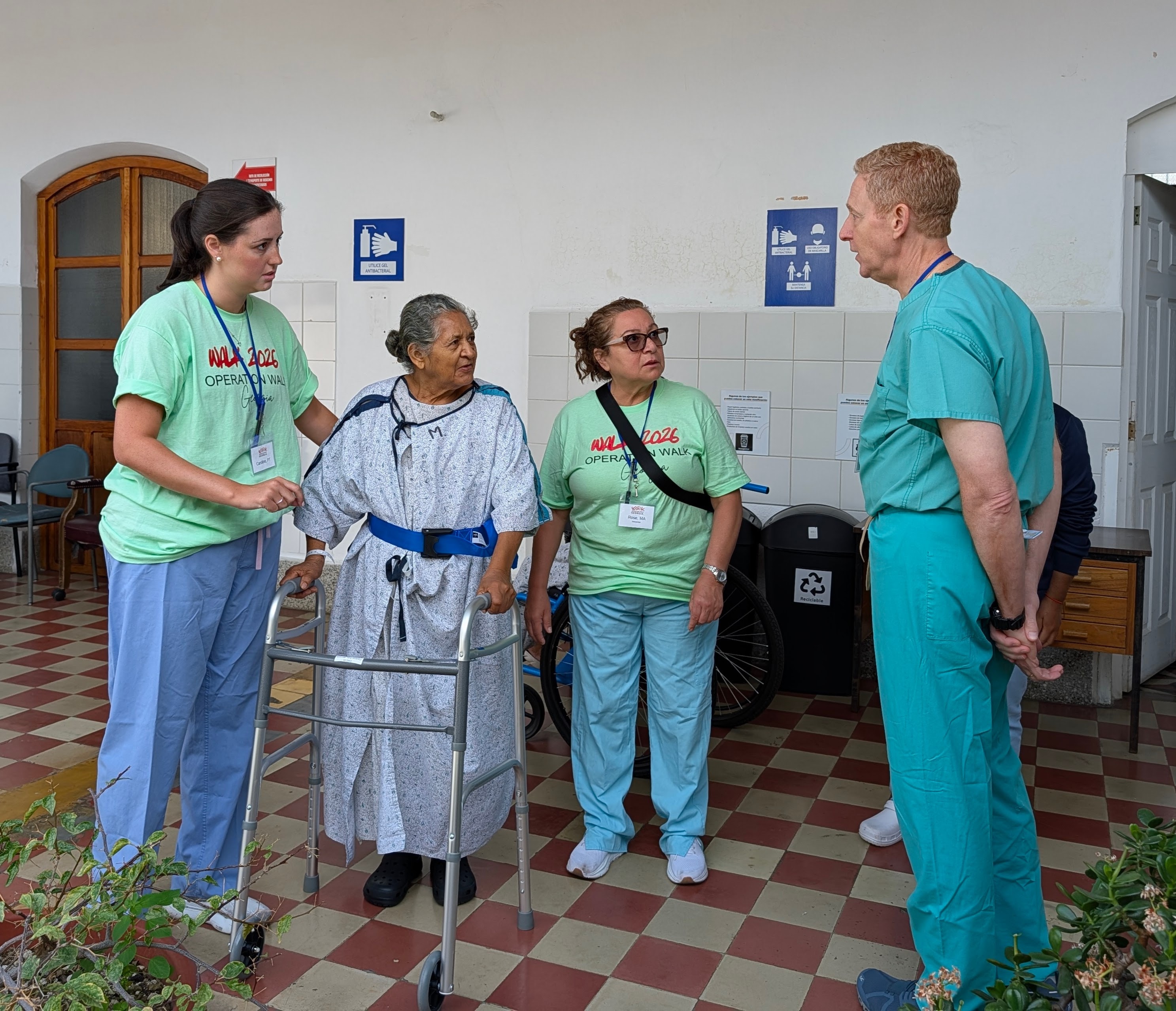 A patient uses a walker with team members beside her in the hospital corridor.